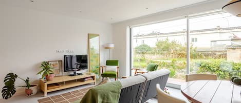 Un salon moderne doté d’un téléviseur à écran plat, d’un canapé, d’une table à manger et offrant une vue sur un jardin grâce à de grandes fenêtres.