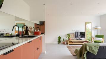 Eine moderne Küche mit einer großen Kochinsel, ein Essbereich mit Fernseher und Sitzgelegenheiten sowie ein Wohnzimmer mit Sofa und grünem Sessel.