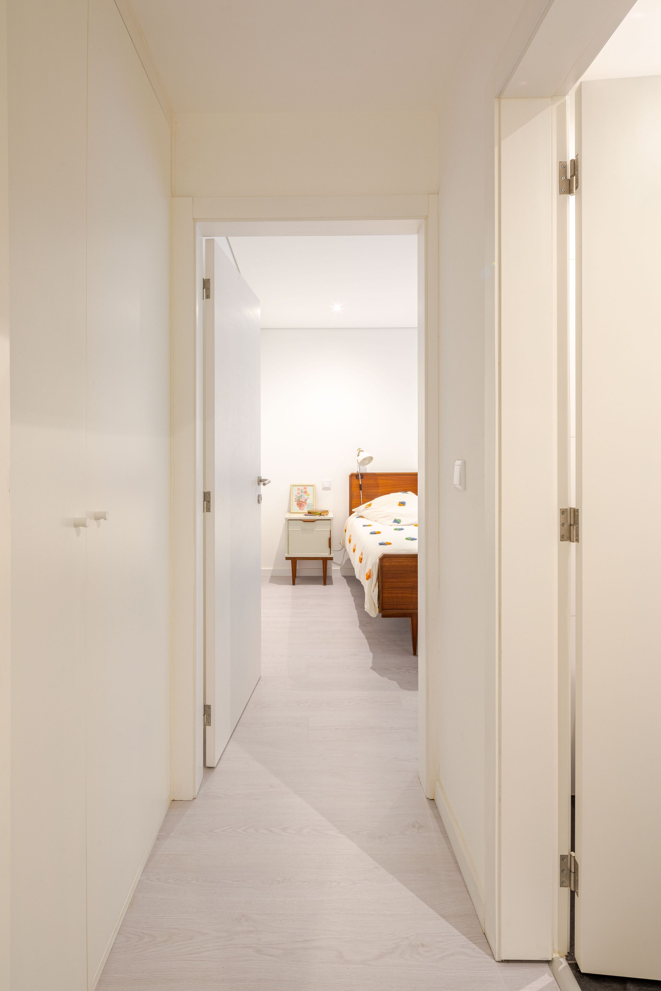 A narrow hallway leading to a bedroom with a bed, bedside table, and a nightstand.