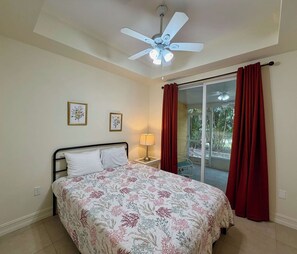 Ein Schlafzimmer mit einem Bett, einem Deckenventilator, einem Fenster mit Vorhängen und Blick auf Grünfläche.
