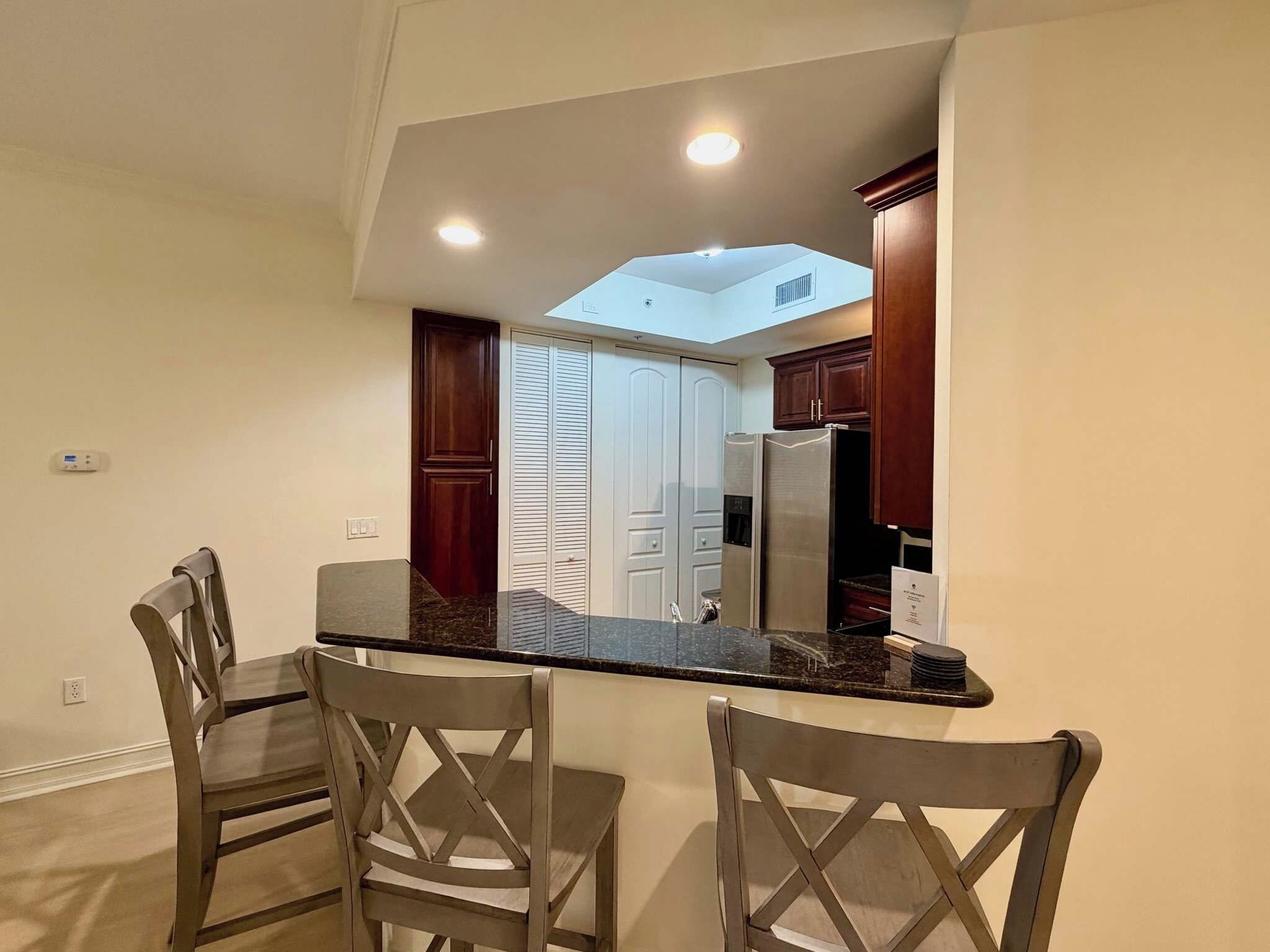 Una cocina con encimera oscura, sillas de madera y vistas a un pasillo.