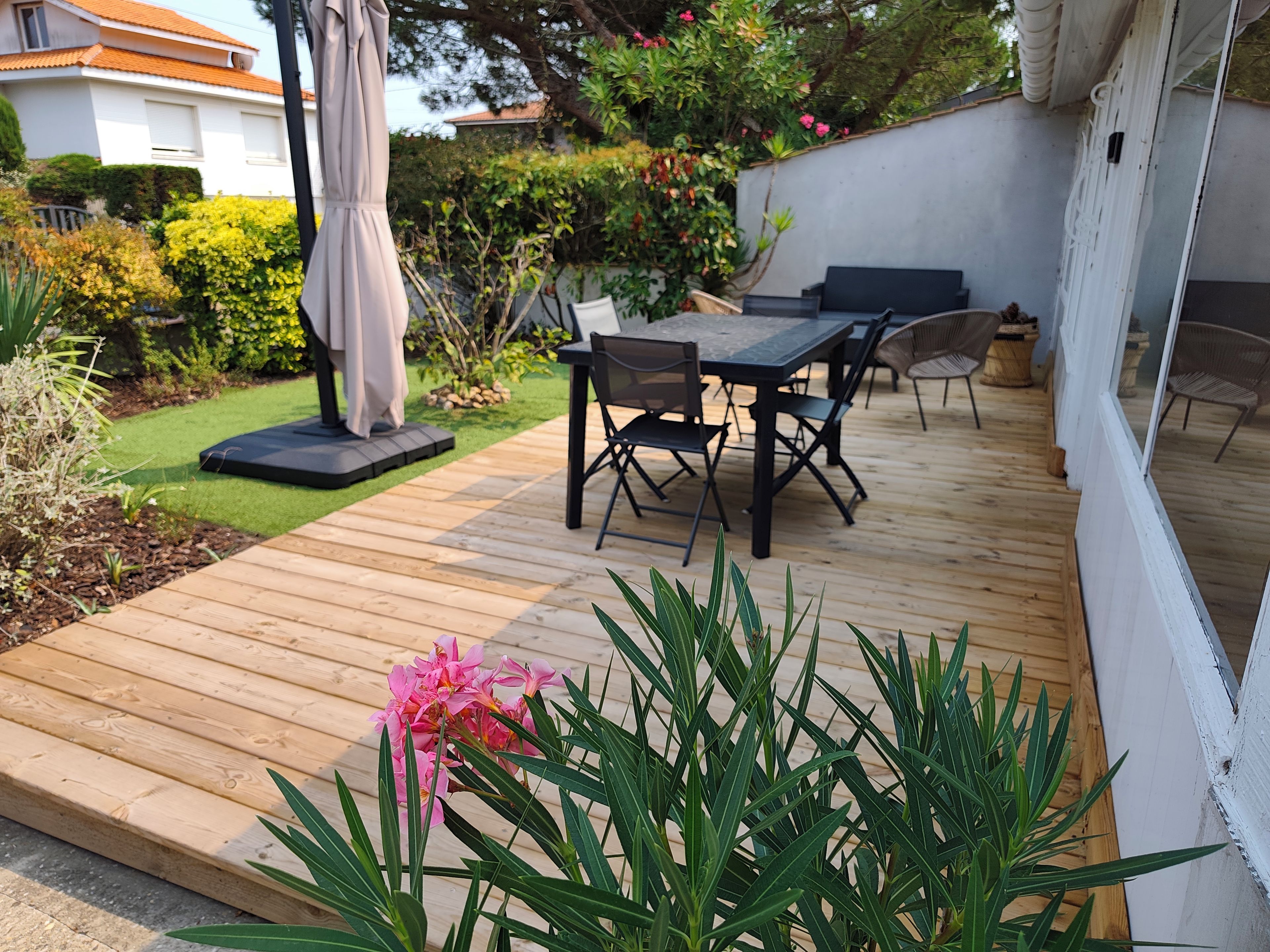 Une terrasse en bois avec une table et des chaises, entourée de verdure et bordée d’un mur blanc.