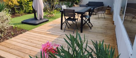 A wooden deck with a dining table and chairs, surrounded by greenery and a white wall.