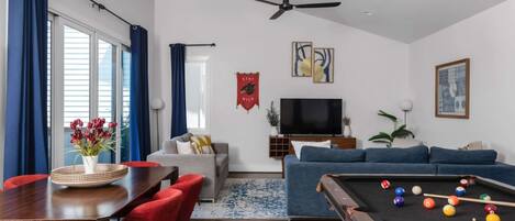 Uma sala de estar com mesa de bilhar, sofá, televisão e um espaço de jantar com cadeiras e mesa.