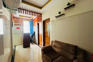 Una habitación con un sofá marrón, un ventilador de techo, una puerta de madera y un estante con una planta.