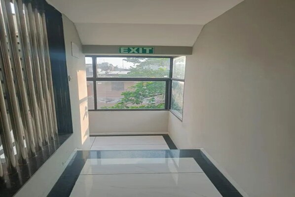 A hallway with an exit sign, a window with a view of buildings and trees, and a tiled floor. - Fabhotel D9 (Indore)