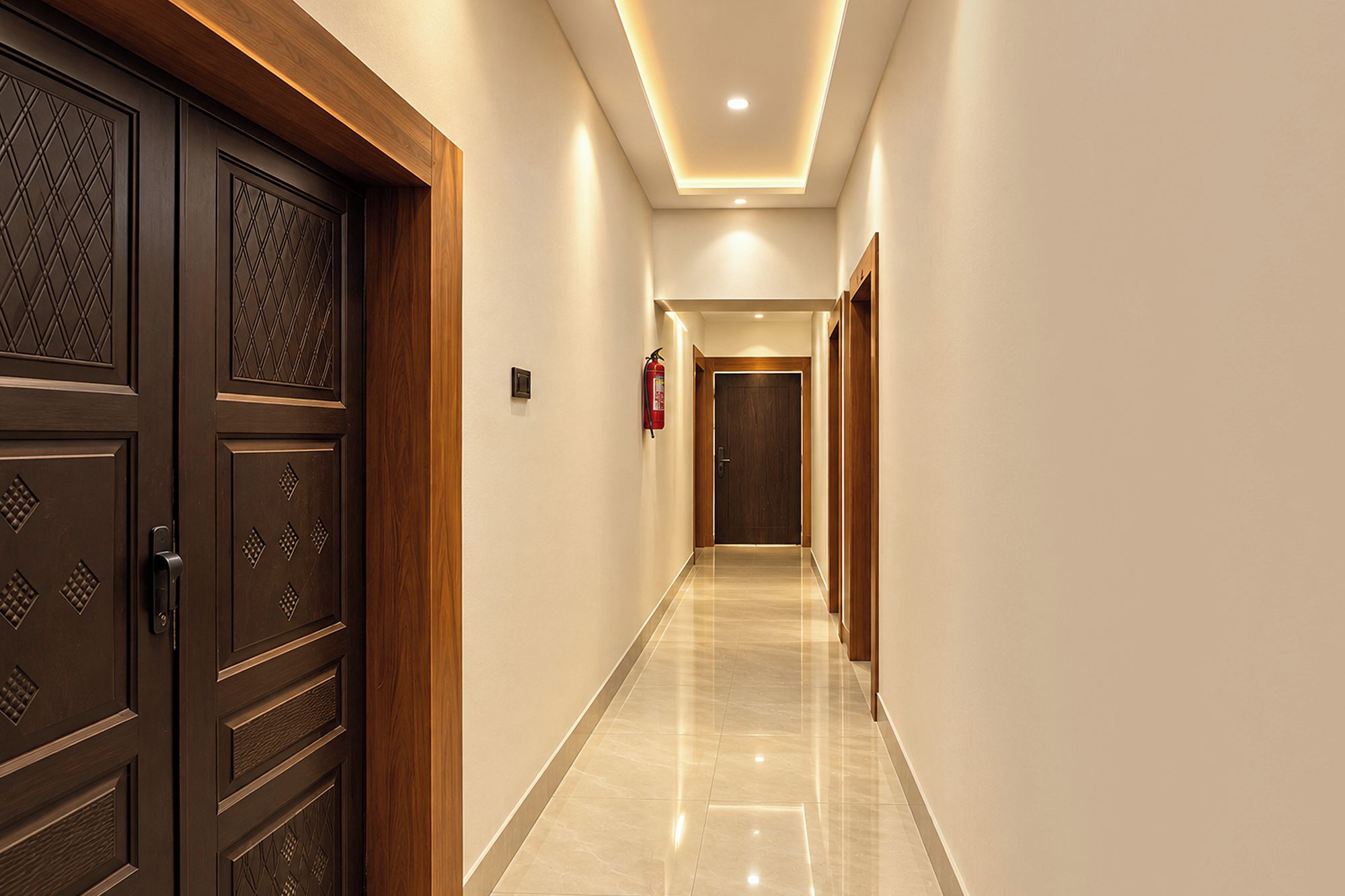 A hallway with a wooden door on the left, a fire extinguisher on the wall, and a door at the end.