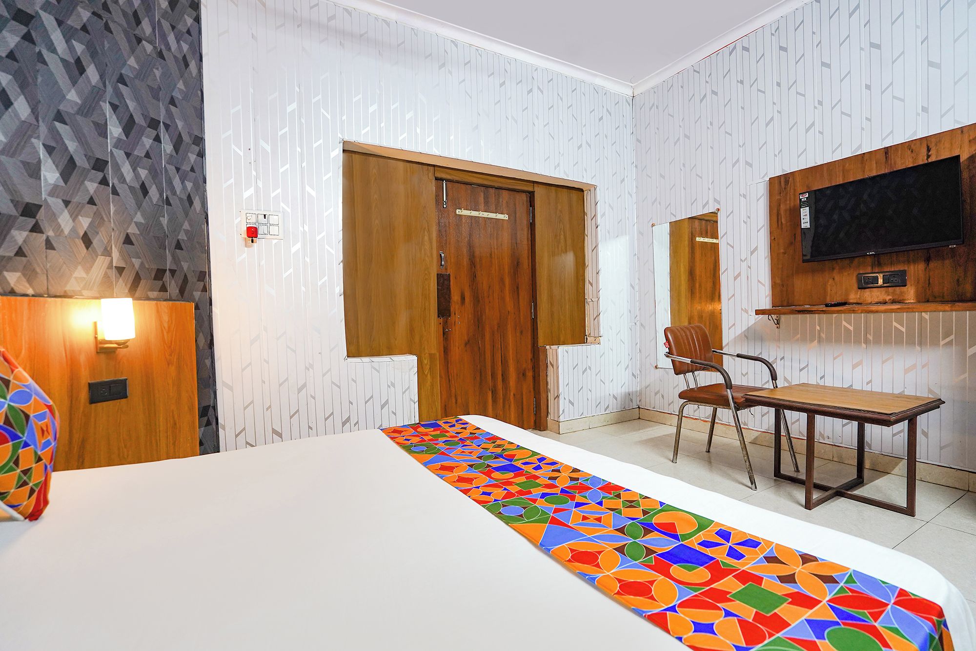 A hotel room with a bed, a desk, a chair, a TV mounted on the wall, and a patterned carpet.