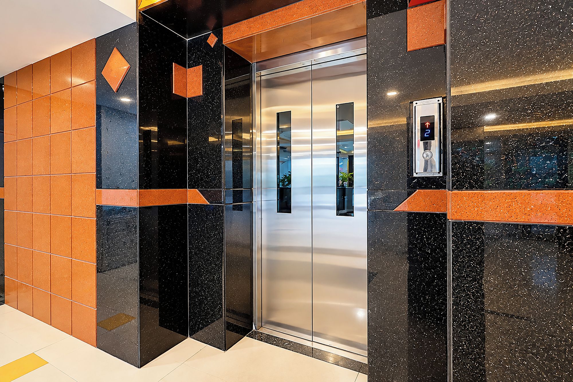 Un ascensor moderno con pantalla digital, rodeado de paredes con azulejos negros y naranjas.
