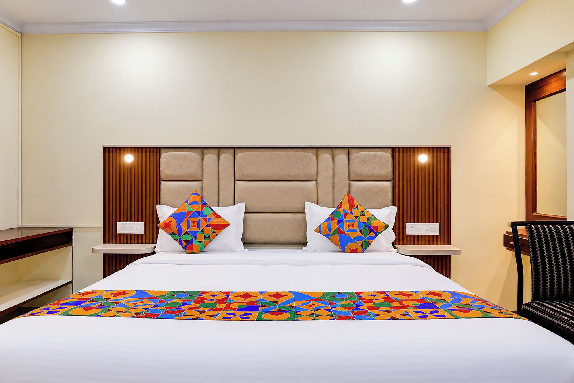 A hotel room with a large bed, two pillows with geometric patterns, a wooden headboard, and a bedside table with a lamp.