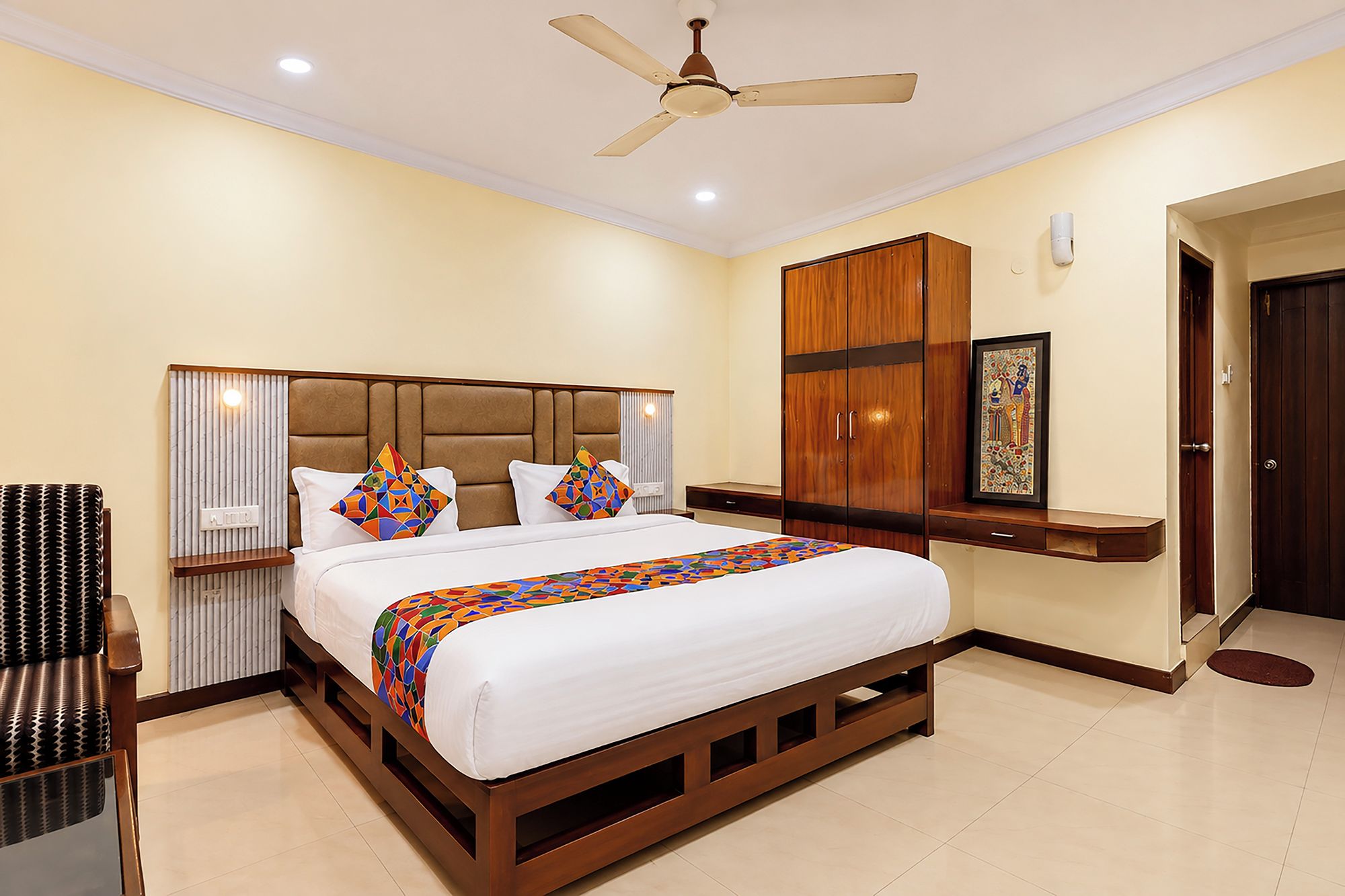 A hotel room with a bed, a ceiling fan, a wooden wardrobe, and a framed picture on the wall.