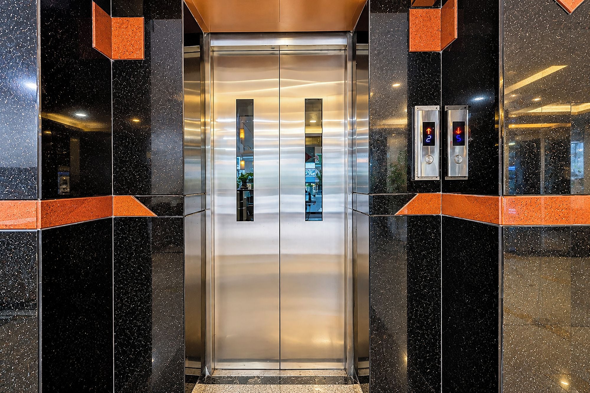 An elevator with a metallic finish, flanked by black and orange-tiled walls, and two illuminated control panels above it.