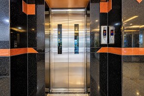 Un ascensor con detalles metálicos, flanqueado por paredes cubiertas de baldosas negras y anaranjadas, y dos paneles de control iluminados en la parte superior.