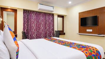 A hotel room with a bed, a television mounted on the wall, an air conditioning unit, a mirror, and a patterned curtain.