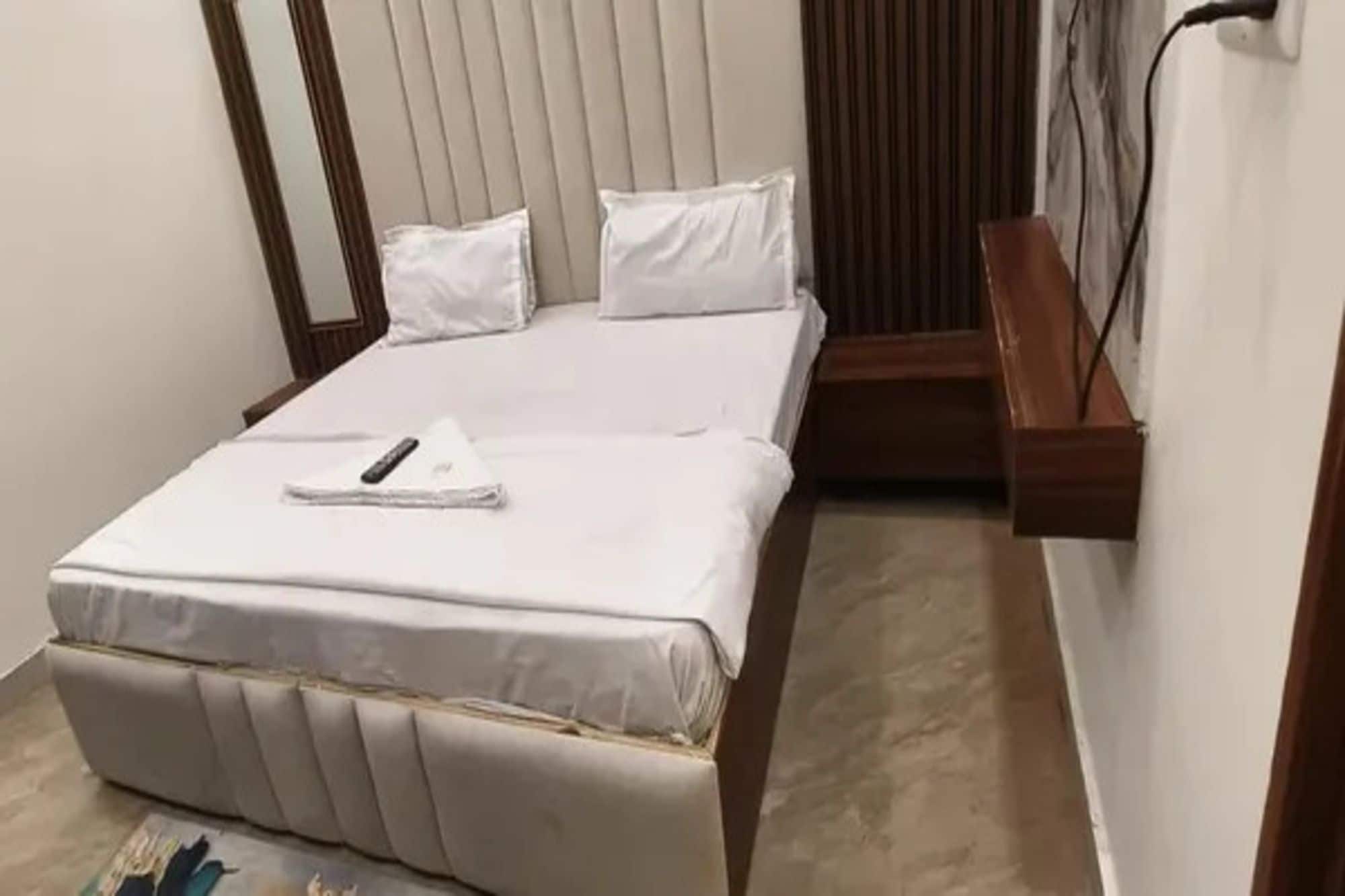 Cama arrumada com controle remoto, um banco de madeira encostado na parede e uma janela com persianas.