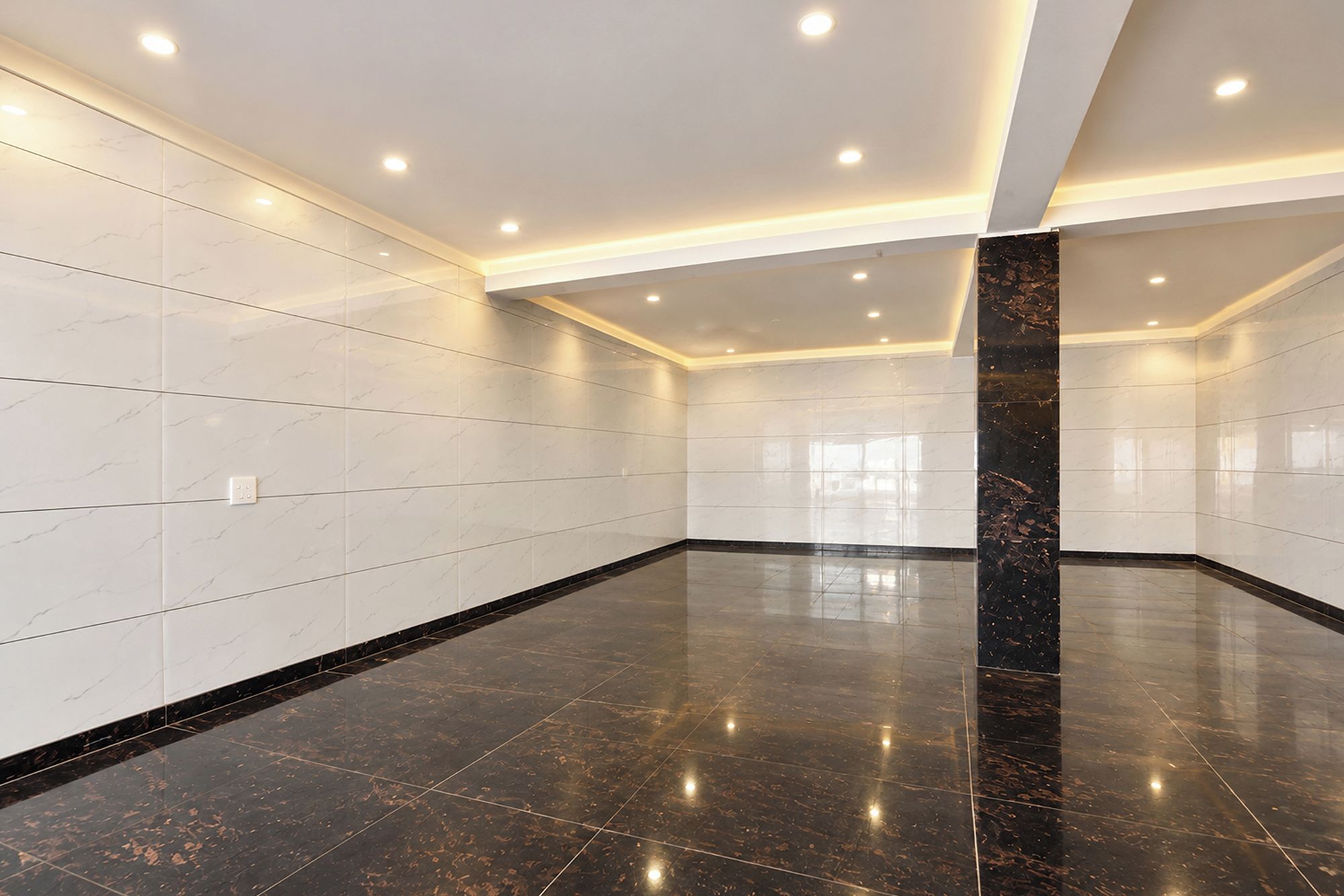 Una habitación amplia con paredes de azulejos blancos, una columna de mármol negro pulido e iluminación empotrada en el techo.