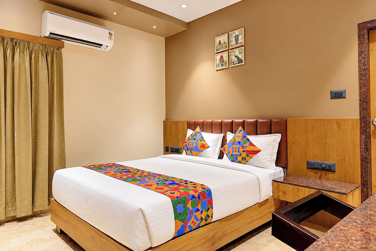 Habitación de hotel con cama, cabecero de madera, mesita de noche, unidad de aire acondicionado, ventana con cortinas y una pared decorada con cuadros.