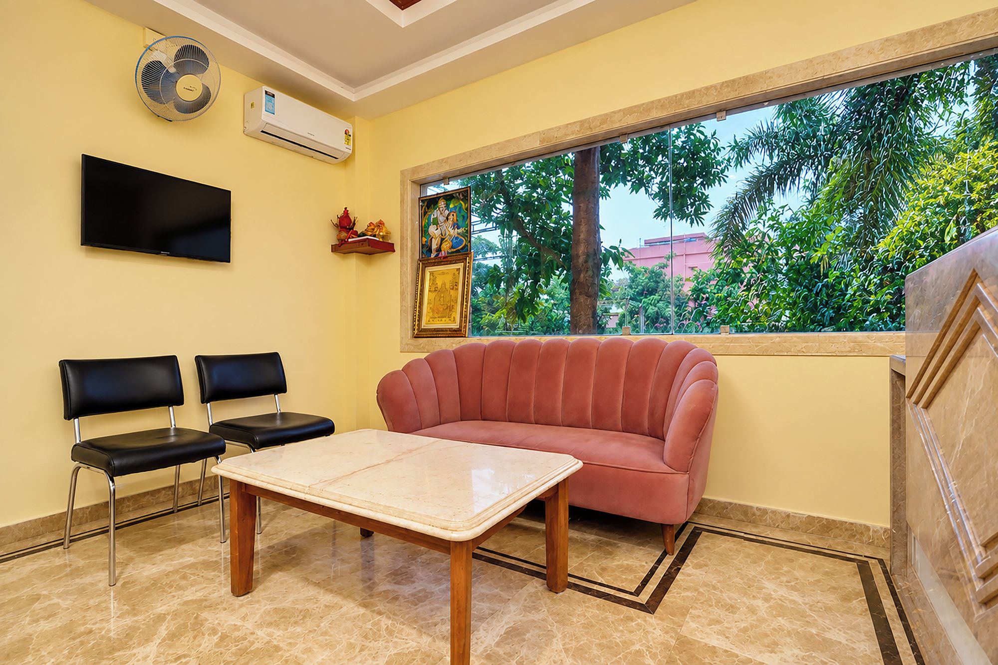 Una habitación con un sofá rojo, una mesa, dos sillas, un televisor, aire acondicionado y una ventana con vistas a árboles.