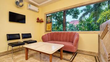 Una habitación con un sofá rojo, una mesa, dos sillas, un televisor, aire acondicionado y una ventana con vistas a árboles.