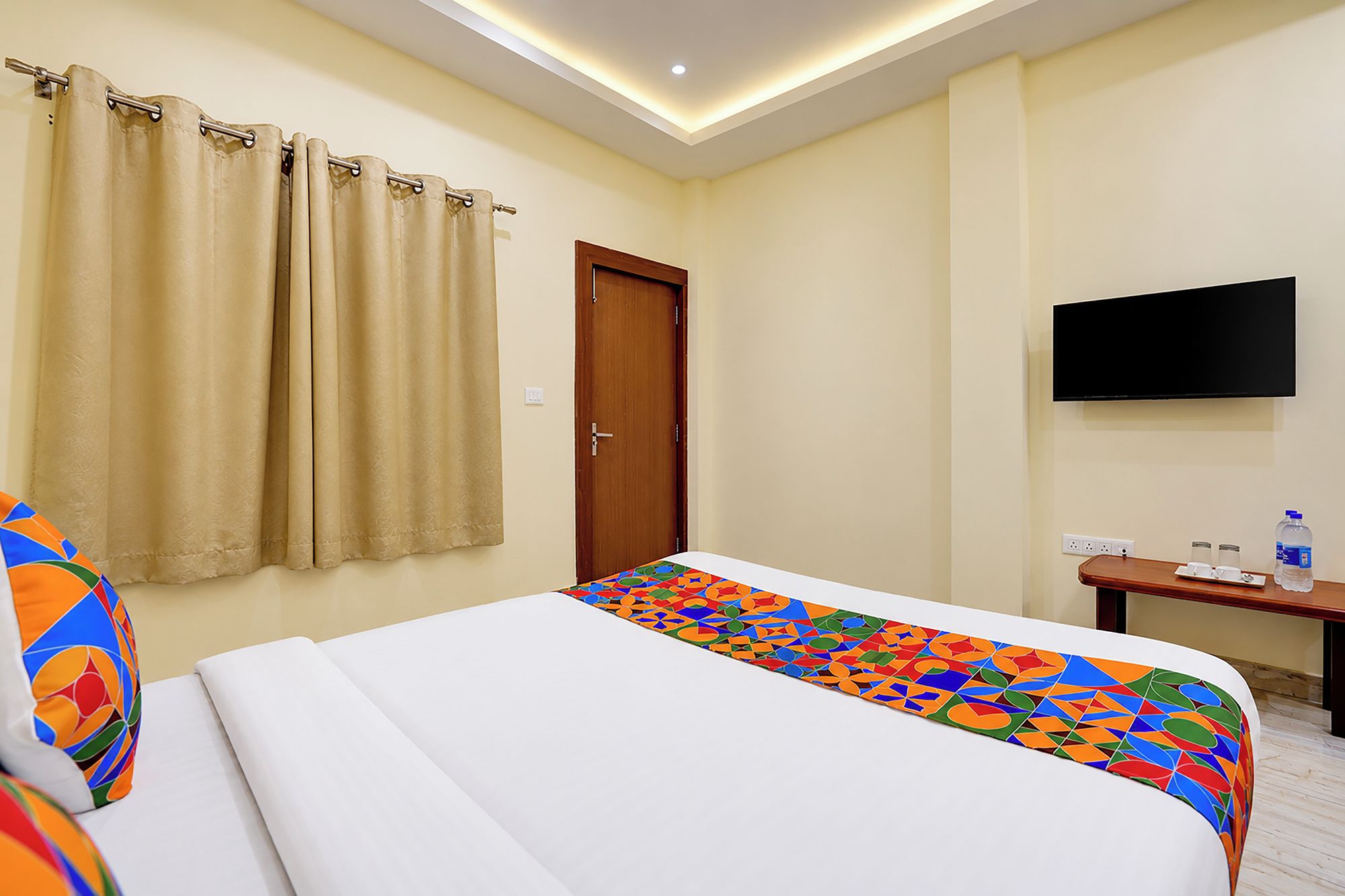 A hotel room with a bed, a television mounted on the wall, a small table with a water bottle, and a door.