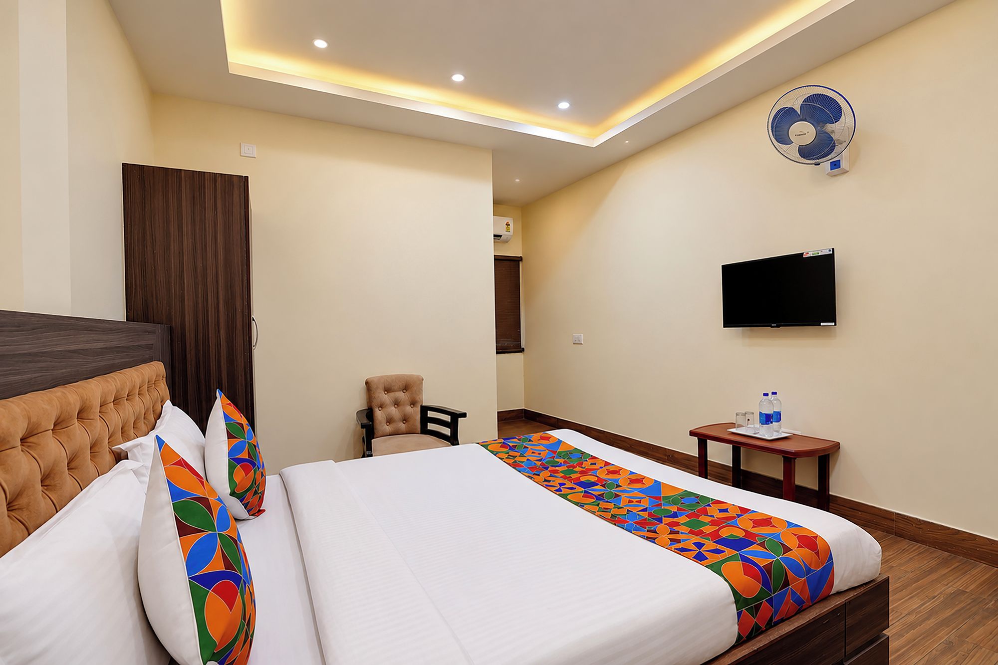 A hotel room with a bed, a chair, a small table with water bottles, a television, and a ceiling fan.