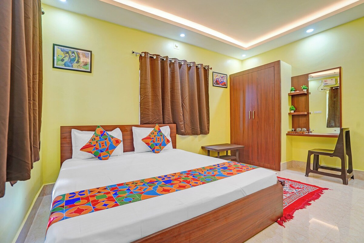 A hotel room with a bed, curtains, a wooden wardrobe, a small table, a mirror, and a red rug.