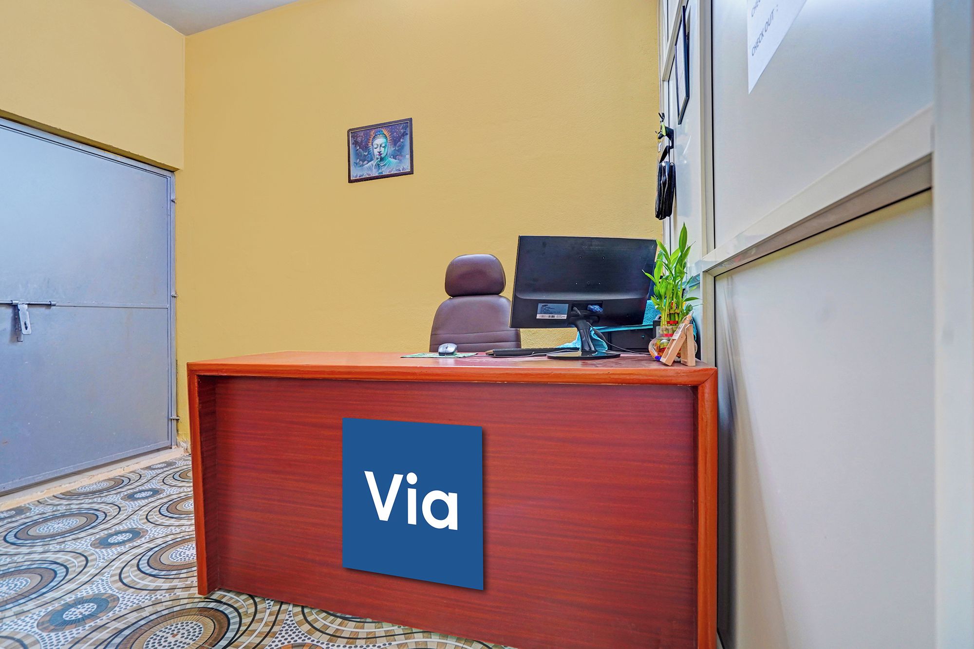 Un mostrador de recepción con un cartel que dice 'Via', un monitor de computadora, una silla, una pequeña estatua de Buda y una planta en maceta.