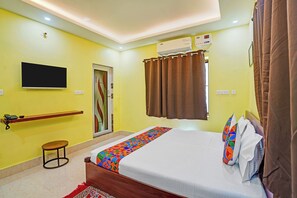A hotel room with a bed, a TV, an air conditioning unit, a curtain, and a small stool. - FabHotel Equinox Inn (Bhubaneshwar)