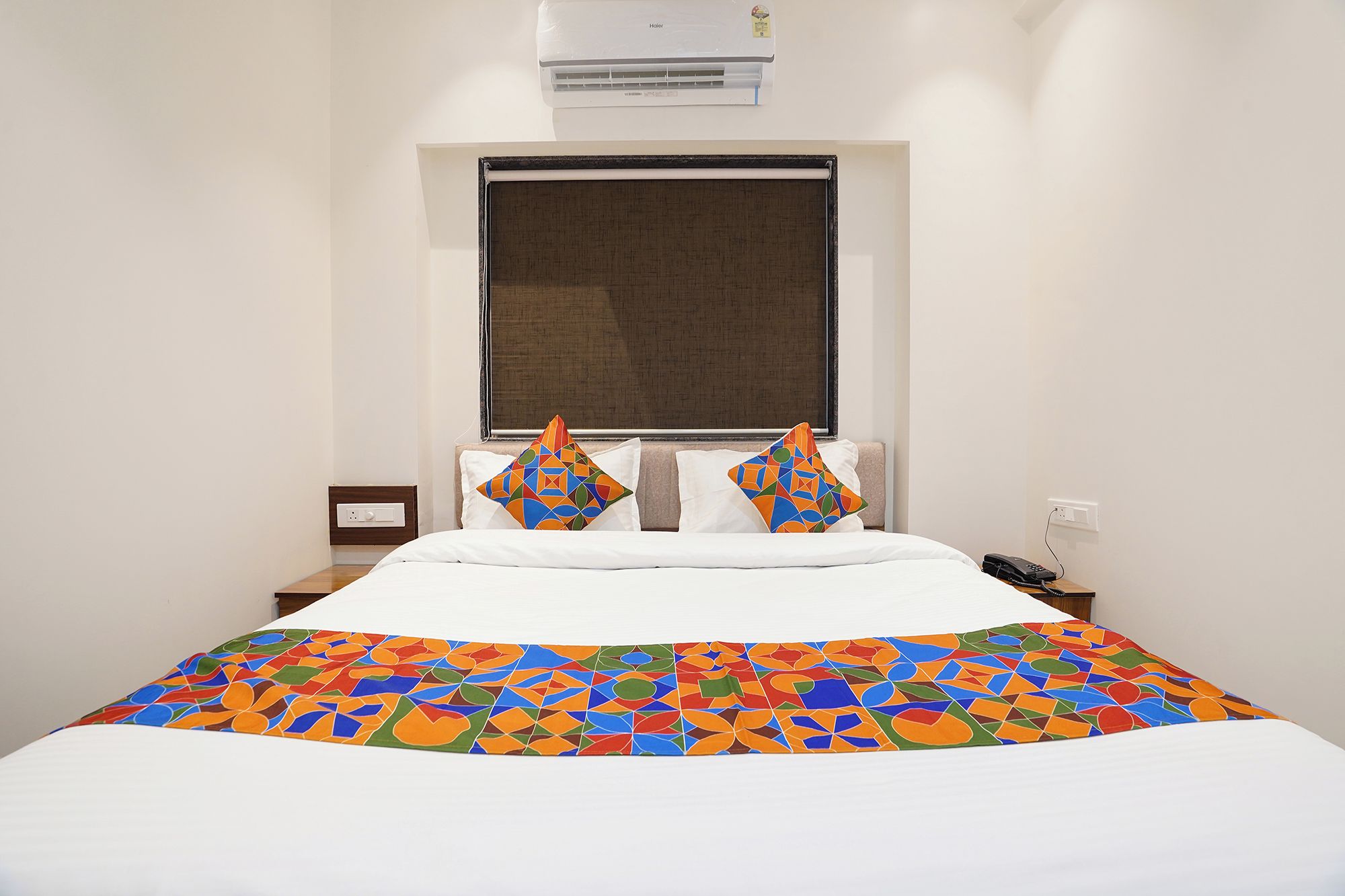 Habitación de hotel con cama, ventana con persiana, mesita de noche con teléfono y un equipo de aire acondicionado en la pared.
