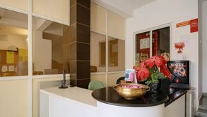 Bancada de recepção com balcão preto, máquina de café Nescafé e um arranjo de flores cor-de-rosa.