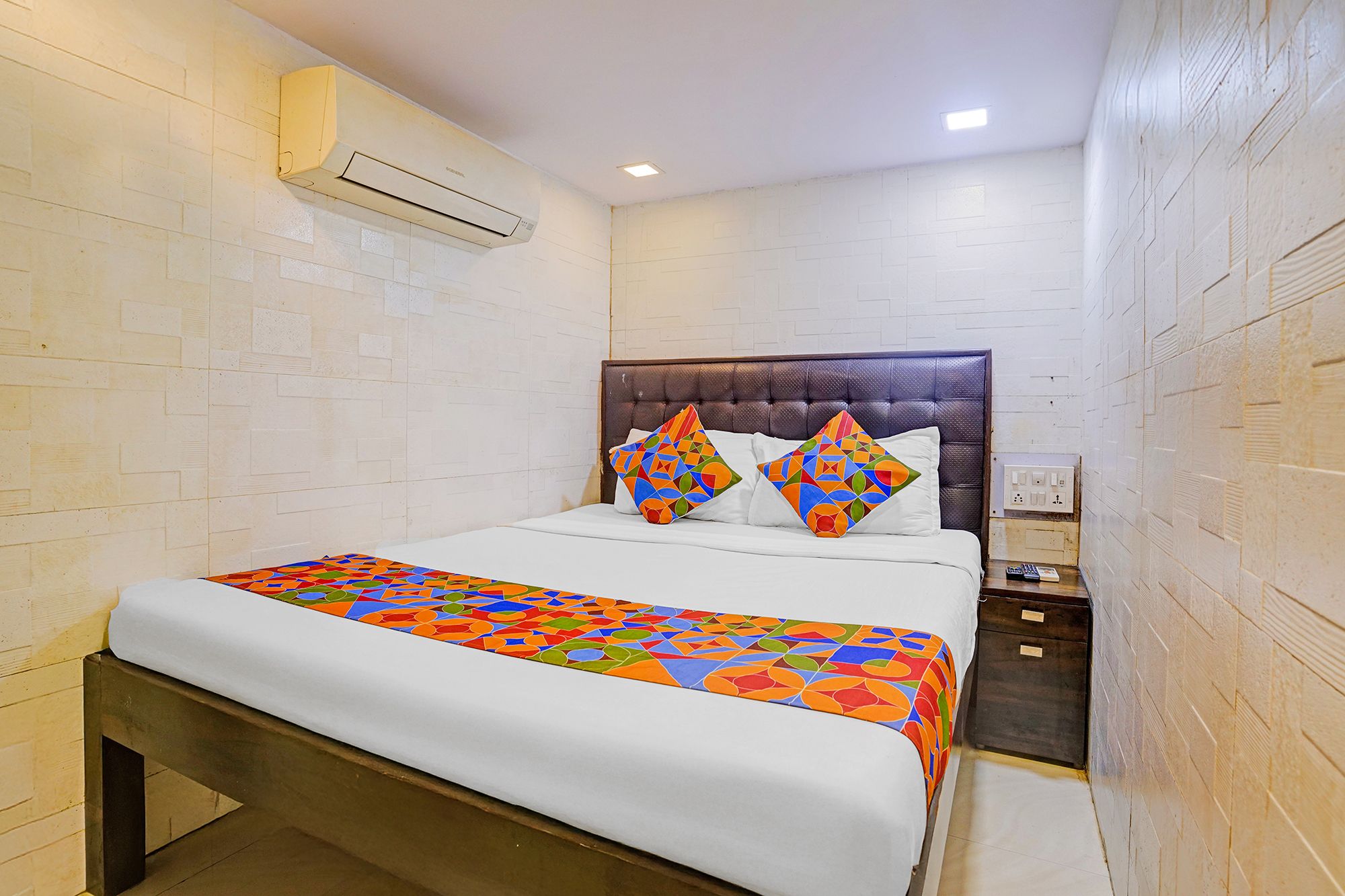 A hotel room with a bed, a nightstand, an air conditioning unit, and a wall-mounted electrical outlet.