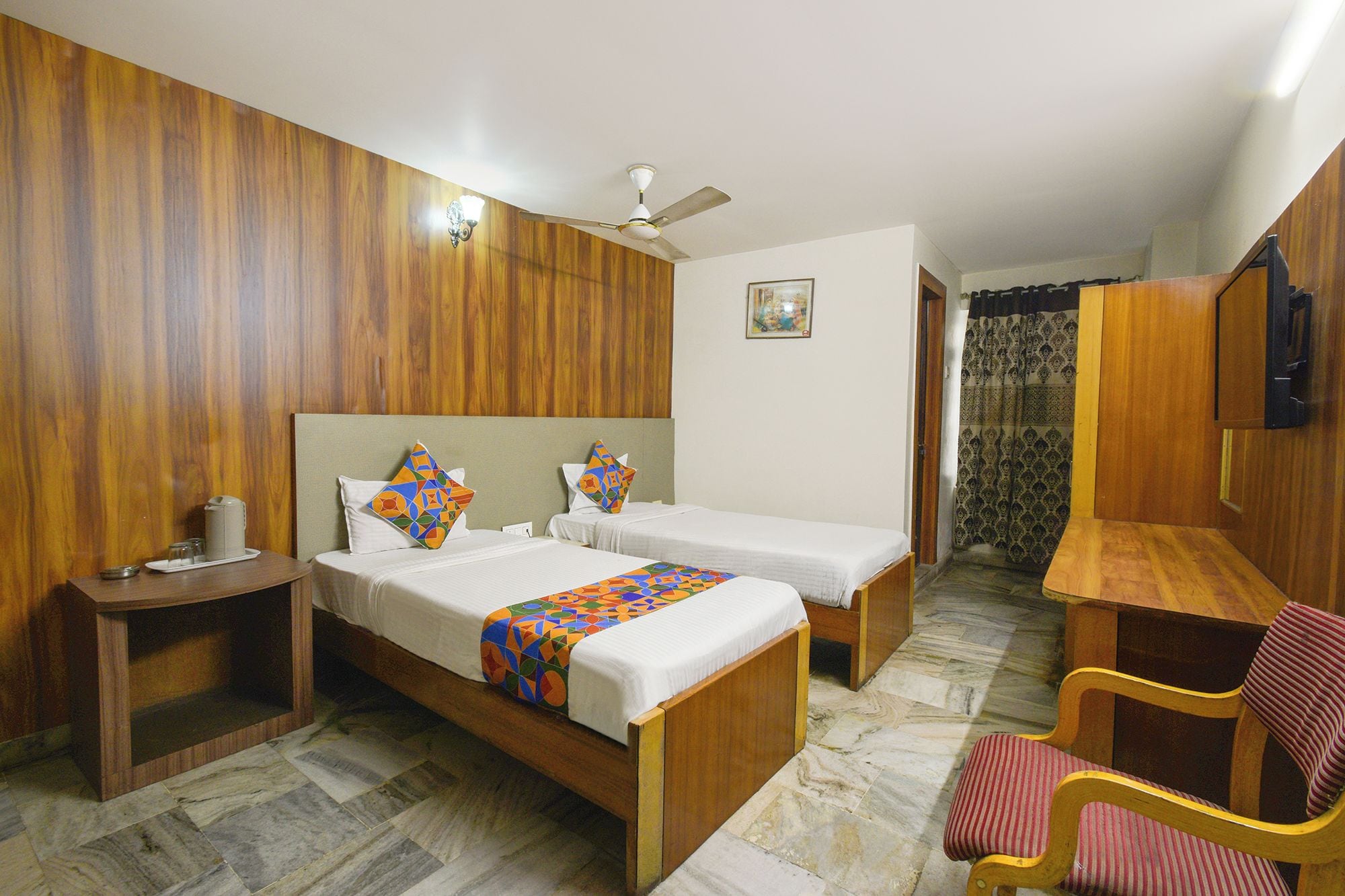 Habitación de hotel con dos camas individuales, cabecera de madera, mesita de noche con lámpara, una silla y un televisor en la pared.