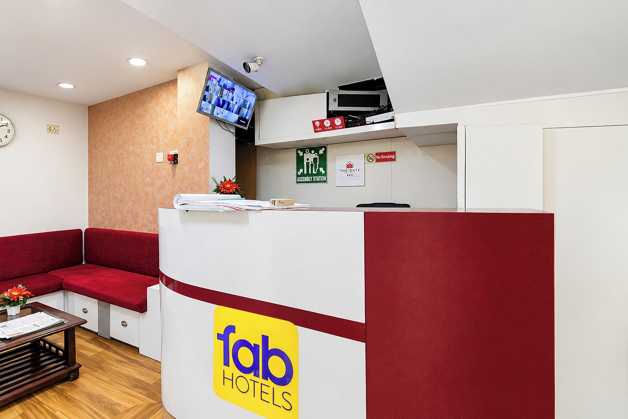 Zona de recepción de hotel con un mostrador rojo y blanco, un reloj y un televisor montado en la pared.