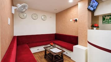 A reception area with a red sofa, a small wooden table with a newspaper, a wall-mounted fan, and a television screen displaying multiple images.