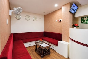 A reception area with a red sofa, a small wooden table with a newspaper, a wall-mounted fan, and a television screen displaying multiple images.