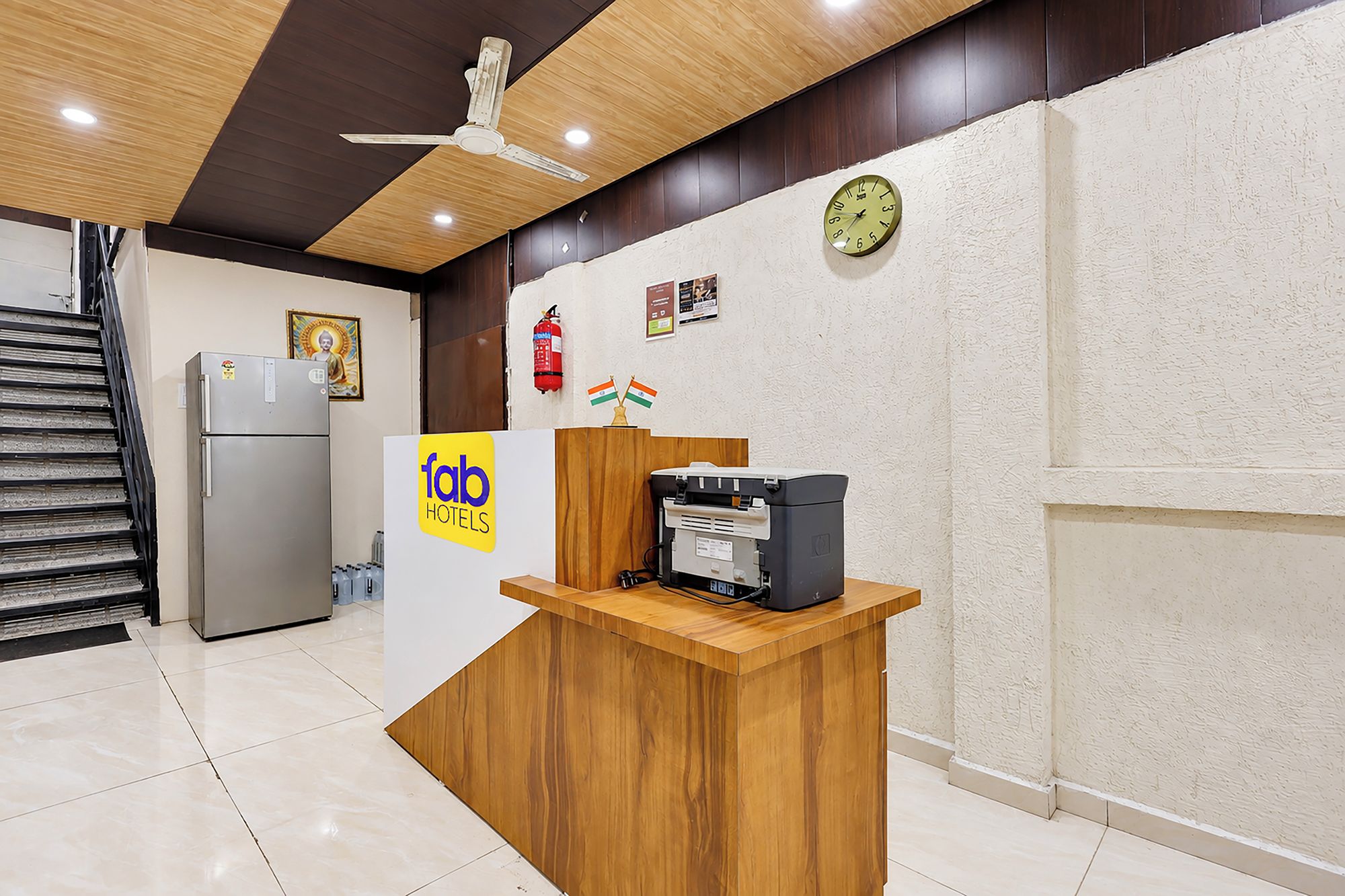 A hotel reception desk with a wooden counter, a refrigerator, a clock, and a ceiling fan.
