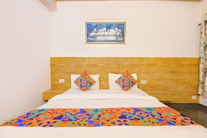 Quarto de hotel com cabeceira de madeira, duas almofadas decorativas, um quadro emoldurado acima da cama e um edredom estampado.