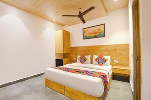 A bedroom with a wooden headboard, a bed with white bedding and colorful geometric pillows, a ceiling fan, and a painting on the wall. - FabHotel Orchid Blu (New Delhi)