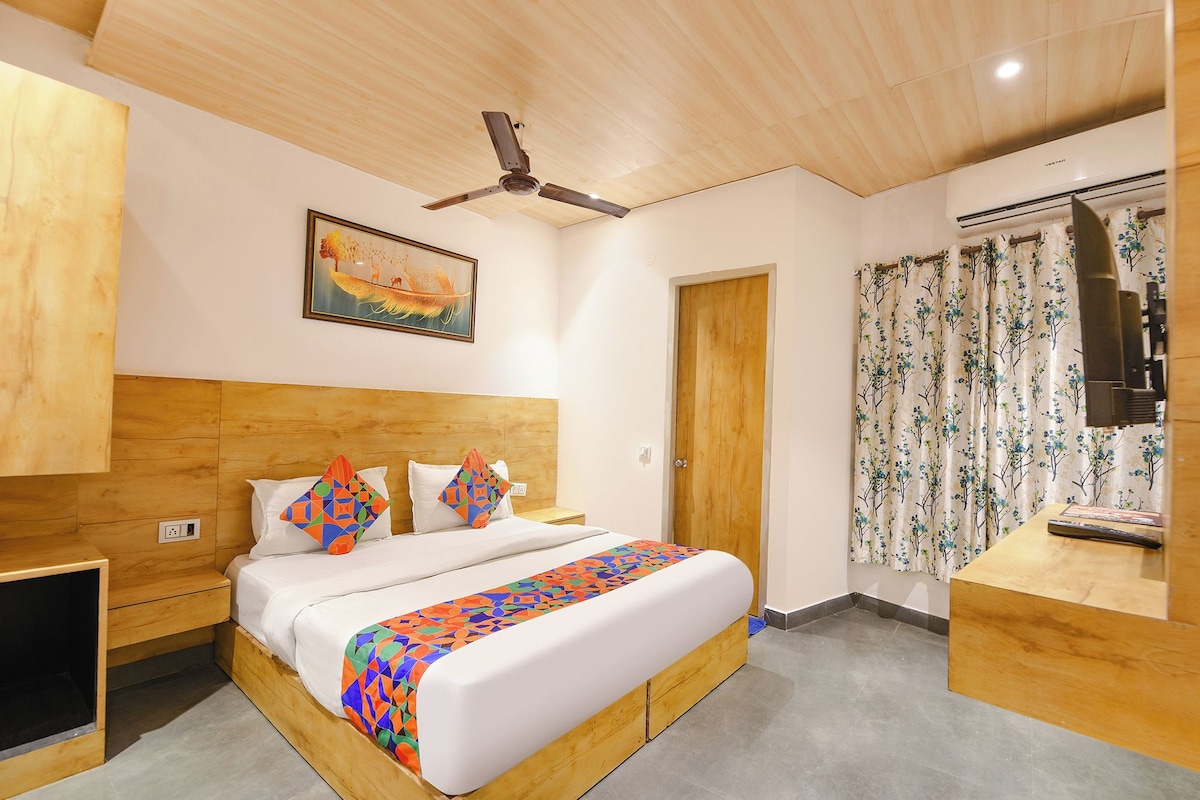 A bedroom with a wooden headboard, a bed with colorful pillows, a ceiling fan, a painting, a door, and a window with floral curtains.