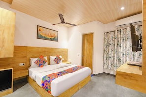 A bedroom with a wooden headboard, a bed with colorful pillows, a ceiling fan, a painting, a door, and a window with floral curtains. - Fabhotel Orchid Blu (New Delhi)