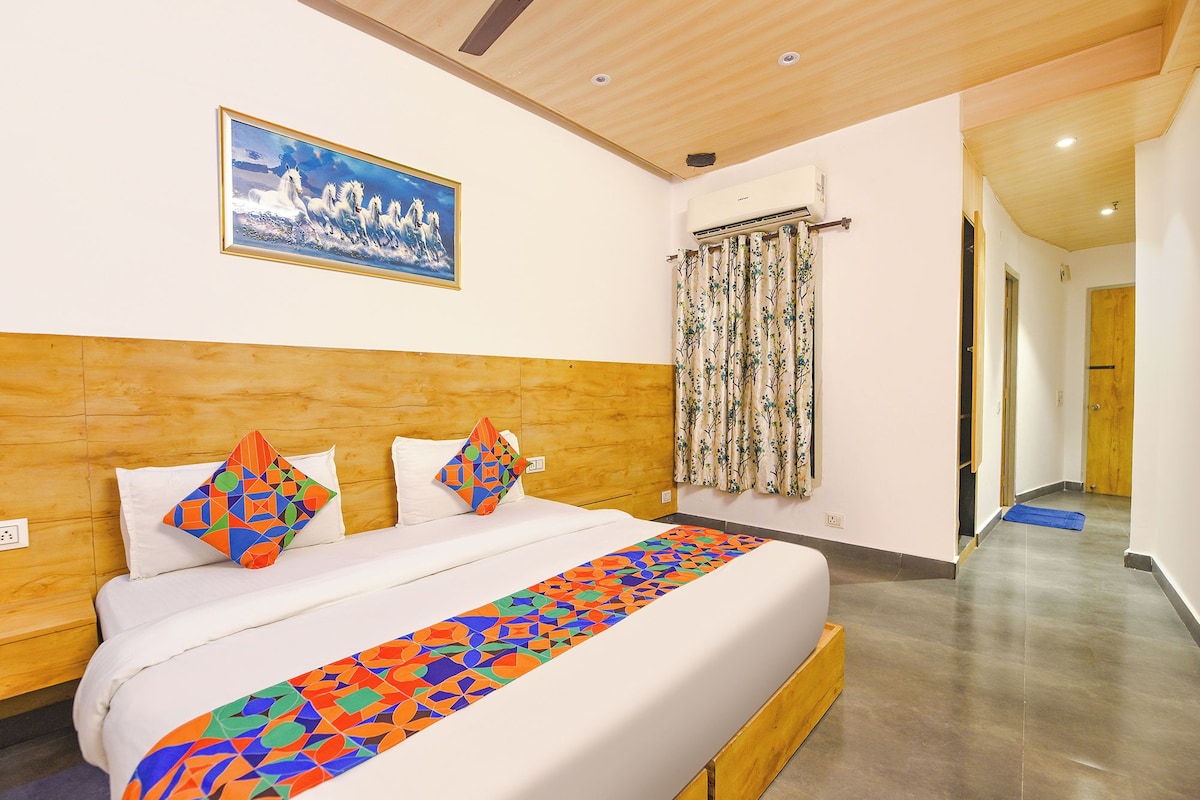 A hotel room with a wooden headboard, a bed with colorful geometric pillows, a large painting of horses, an air conditioning unit, and a patterned curtain.