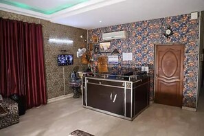 Une pièce avec un papier peint à motifs, une porte en bois, un téléviseur fixé au mur et un bureau avec des tiroirs.