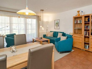 Ein Wohnzimmer mit einer Couch, einem Couchtisch, einem Esstisch mit Stühlen, einem Bücherregal und einem Fenster mit Vorhängen.