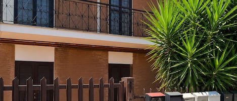 Un edificio residencial con entrada protegida por una reja, un balcón con barandilla de hierro forjado y una planta grande al frente.