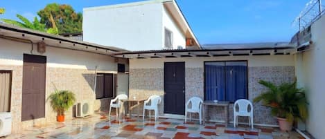 A courtyard with a tiled floor, a white building, and a covered seating area with chairs.