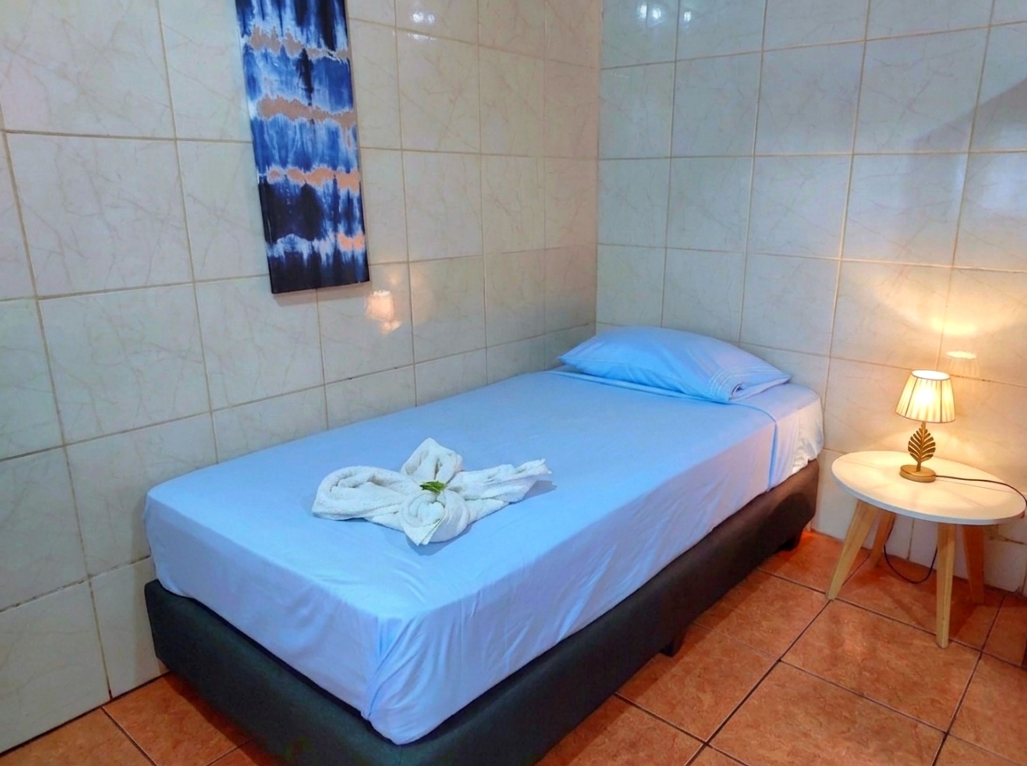 Un lit simple avec un matelas bleu et une couverture blanche, une serviette sur le lit, une petite table de chevet avec une lampe et un tableau au mur.