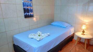 Un lit simple avec un matelas bleu et une couverture blanche, une serviette sur le lit, une petite table de chevet avec une lampe et un tableau au mur.