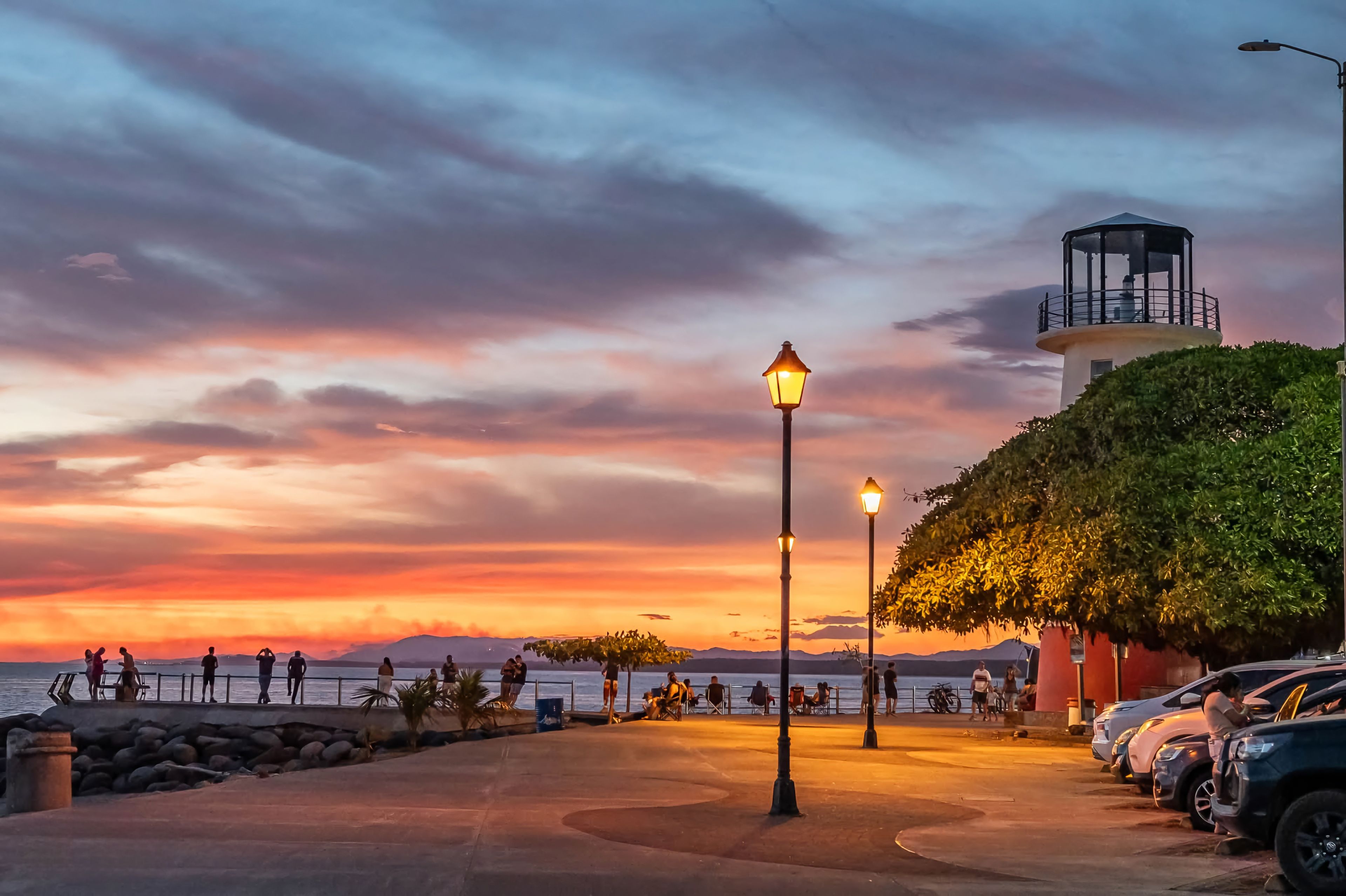 Un paseo marítimo con un faro, farolas y coches estacionados.