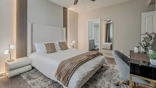 Une chambre moderne comprenant un lit, des tables de chevet, un bureau, une chaise, un ventilateur de plafond et une salle de bain visible à travers une porte ouverte.