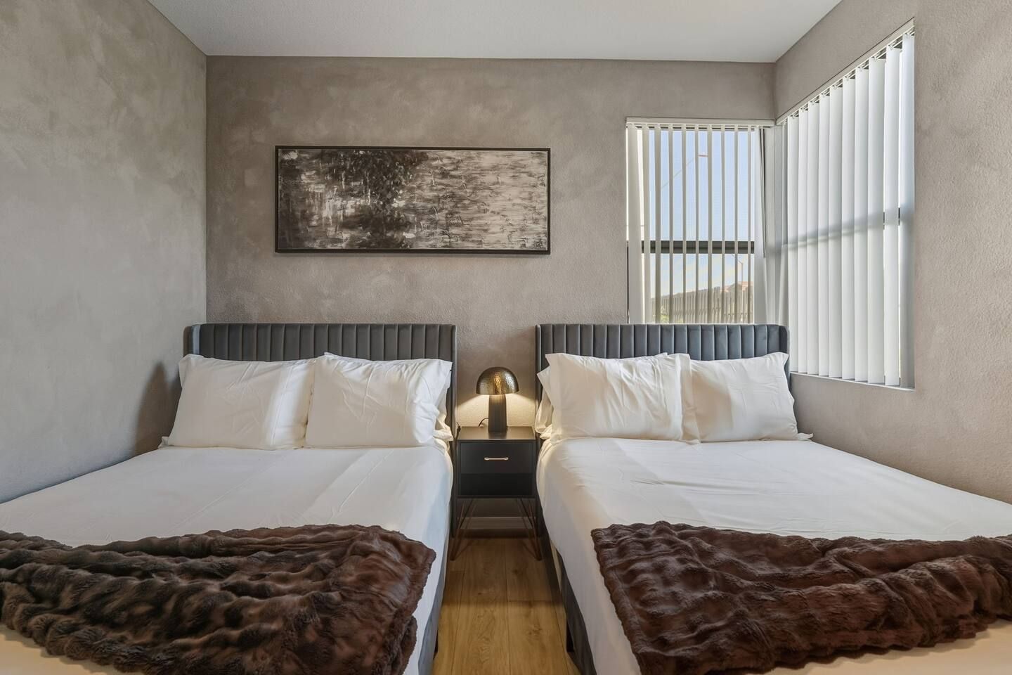 A hotel room with two beds, a nightstand, a lamp, a painting on the wall, and a window with blinds.